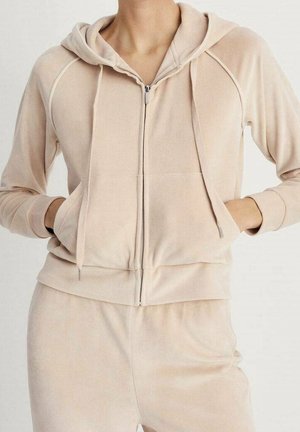 Person iført en beige hættetrøje med lynlås og matchende joggingbukser med hænderne i forlommerne, stående foran en ensfarvet baggrund.