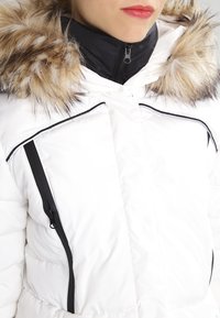 Bílá péřová bunda s odnímatelným límcem z umělé kožešiny, černým zipem a kontrastními černými detaily na zipy a švech.