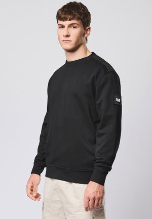 Joven con sudadera negra de manga larga con parche en la manga y pantalones beige, de pie frente a un fondo liso gris claro.
