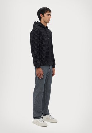Sort hættejakke med et struktureret geometrisk mønster, der har snøre i hætten og en frontlomme, parret med grå denim jeans og hvide sneakers.