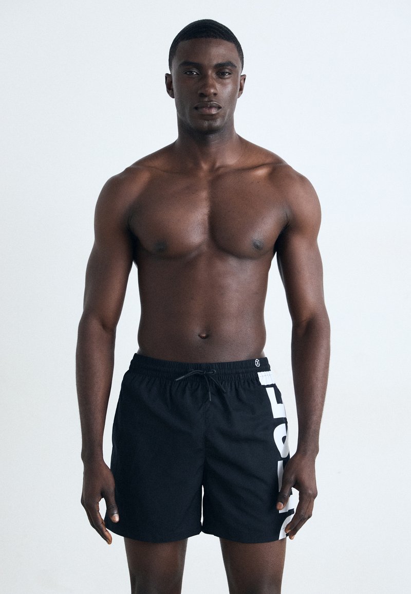 Black swim shorts with a drawstring waistband and large white lettering on the side. The material appears lightweight and smooth.