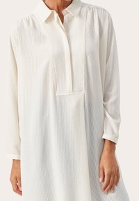 Paita long-sleeve longgar berwarna putih dengan kain bertekstur, dilengkapi kerah, placket butang, dan perincian jahitan di panel depan.