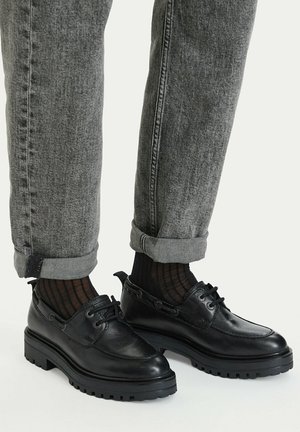 Persona que lleva zapatos negros de cuero con cordones, calcetines oscuros con estampado y vaqueros grises con el dobladillo vuelto, sobre un fondo claro y liso.