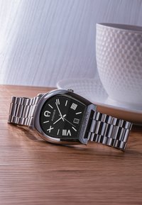 Stříbrné nerezové hodinky s černým obdélníkovým ciferníkem, bílými indexy a zobrazením data. Umístěny vedle bílé texturované hrnku.