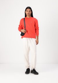 Felpa rossa con polsini a coste, abbinata a pantaloni della tuta crema. Una borsa a tracolla nera e sneaker nere completano l'outfit.