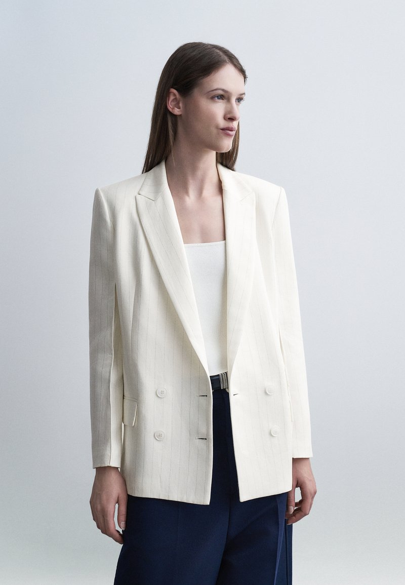 Femme portant un blazer croisé à fines rayures blanches sur un haut blanc et un pantalon bleu marine, debout devant un fond clair uni.