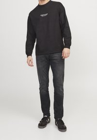 Camisa negra de manga larga con logo blanco en el pecho, combinada con vaqueros oscuros ajustados y zapatillas negras, que exhibe un look casual.