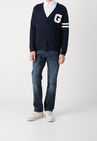 Marinblå cardigan med V-hals, knappar, två framfickor och ett stort vitt 'G' på bröstet. Buren över en vit skjorta och blå jeans.