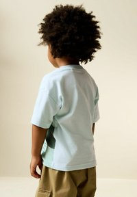 Tricou din bumbac albastru deschis cu mâneci scurte, cu decolteu rotund și design cu panou lateral, purtat cu pantaloni scurți cargo de culoare bronz.