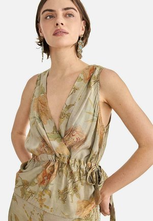Mujer que lleva una blusa sin mangas, cruzada y con estampado floral, con cintura ajustable con cordón, combinada con grandes pendientes colgantes decorativos en azul y dorado.