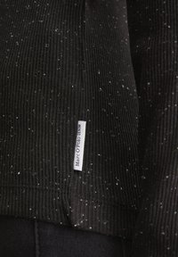 Crna rebrasta tkanina s bijelim pjegama. Uključuje bijelu oznaku s natpisom "Marc O'Polo DENIM." Prikaz u krupnom planu koji ističe teksturu i dizajn.