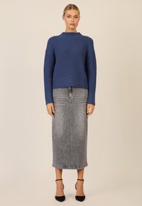 Kobieta ubrana w niebieski sweter z prążkami, szaro-srebrną spódnicę midi z ćwiekami i czarne szpilki z paskiem na kostce, stojąca na jednolitym beżowym tle.