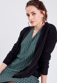 Cardigan en tricot noir sur une robe noire avec un motif floral turquoise. Le cardigan présente une coupe ample avec des poignets côtelés.