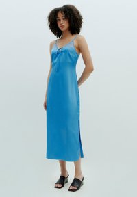 Blaues Satin-Nachthemd mit verstellbaren Spaghetti-Trägern, Spitzenakzenten, seitlichem Schlitz und einer Schleifendetail am Ausschnitt, kombiniert mit schwarzen Mules.