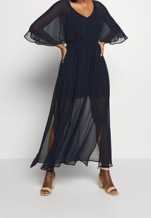 Frau trägt ein marineblaues, transparentes, bodenlanges Kleid mit flatternden Ärmeln und hohen Schlitzen, kombiniert mit beigen Sandaletten mit hohem Absatz.