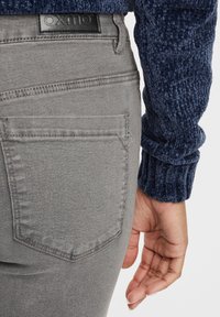 Szürke farmernadrág hátsó zsebbel és fekete márkajelzéssel, texturált tengerészkék pulóverrel párosítva. Testhez simuló fazon, laza stílus.