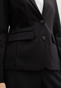 Black blazer featuring a structured design, two front buttons, side pockets, and a smooth texture, paired with a white shirt underneath.