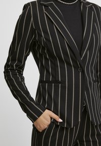 Blazer rayé noir et crème en tissu lisse ; comporte un col à revers cranté, une fermeture à un bouton et des poches avant.