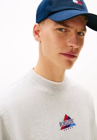 Γκρι φούτερ με στρογγυλή λαιμόκοψη, διακοσμημένο με πολύχρωμο κεντητό λογότυπο. Ναυτικό μπλε καπέλο με επίπεδο γείσο, φορεμένο πάνω σε ανοιχτόχρωμη επιδερμίδα.