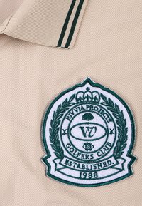 Beiges Polohemd mit einem grünen, gestickten Abzeichen, das einen Lorbeerkranz, den Text "VIA PROJECTS GOLFERS CLUB" und "GEGRÜNDET 1988" zeigt.