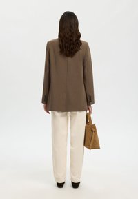 Žena s vlnitými hnědými vlasy, oblečená v hnědém blejzru, krémových kalhotách a černých botách, držící hnědou semišovou kabelku, otočená zády na jednobarevném pozadí.