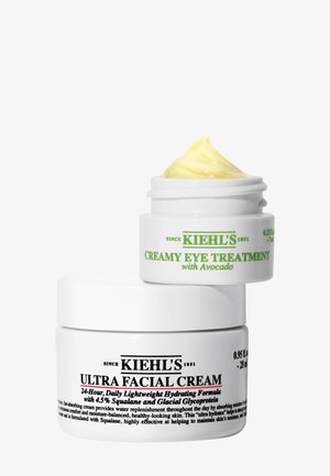 Tarro de crema Ultra Facial de Kiehl’s con un tarro más pequeño abierto de tratamiento cremoso para ojos con aguacate apilados uno encima del otro sobre fondo blanco.