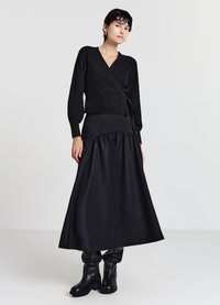 Maglione avvolgente nero con motivo a diamante, abbinato a una lunga gonna nera in satin con orlo arricciato. Stivaletti neri completano l'outfit.