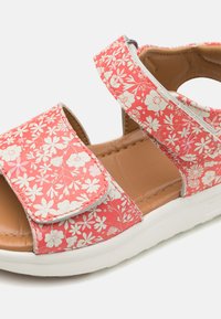 Koralblommönstrad sandal med en bred kardborreband, vadderad sula och vit gummisula. Öppna sidor för ventilation.