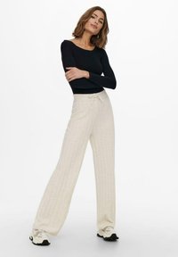 Lange, hellbeige gerippte Hosen mit einem Kordelzugbund, kombiniert mit einem figurbetonten schwarzen Langarmoberteil. Das Model trägt sportliche schwarz-weiße Schuhe.