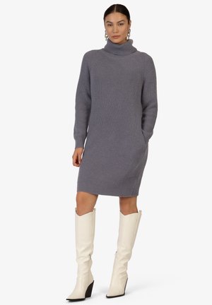 Grau gerippte Strickkleid mit hohem Rollkragen, langen Ärmeln, seitlichen Taschen und knielangen weißen Stiefeln mit spitzer Zehe.