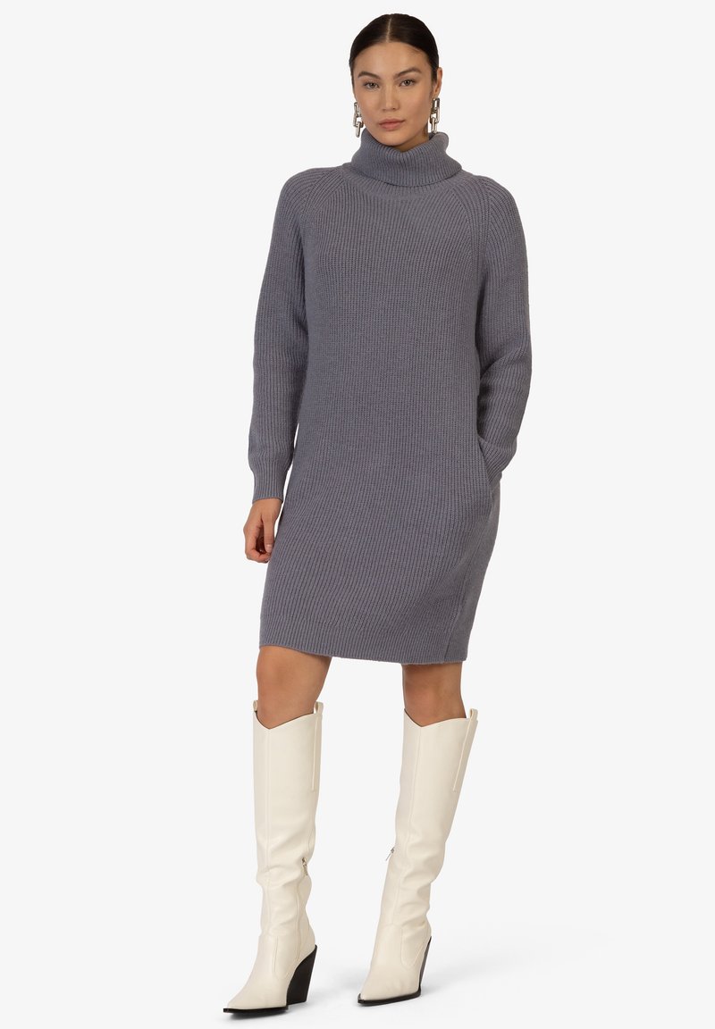 Grau gerippte Strickkleid mit hohem Rollkragen, langen Ärmeln, seitlichen Taschen und knielangen weißen Stiefeln mit spitzer Zehe.