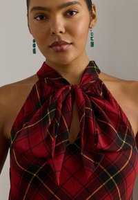 Röd rutig halterneck-topp med rosett vid halsringningen, med svarta och gula linjer. Mjuk tygtextur med en nyckelhålsöppning fram.