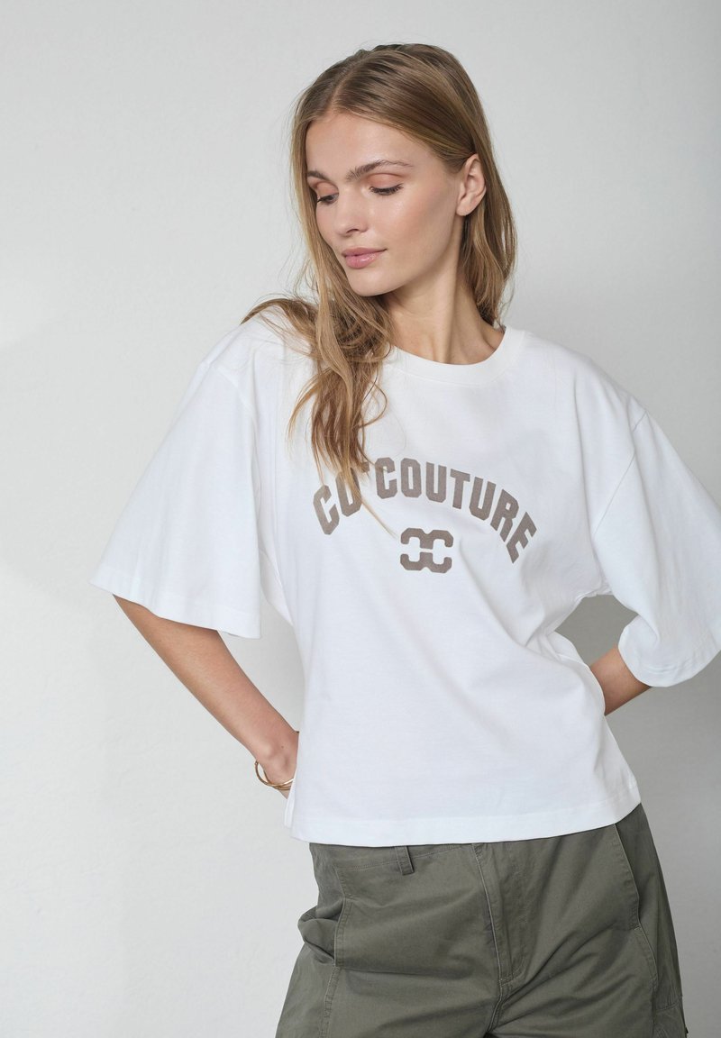 Noor naine pikkade juustega, kellel on seljas valge üle suuruses T-särk, millel on tekst "COCOUTURE" ja logo, kombineeritud roheliste pükstega, poseerib külje pealt.