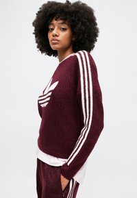 Bordový pletený sveter s bielymi pruhmi značky adidas pozdĺž rukávov a logom na hrudi. Voľný strih s rebrovanou textúrou.