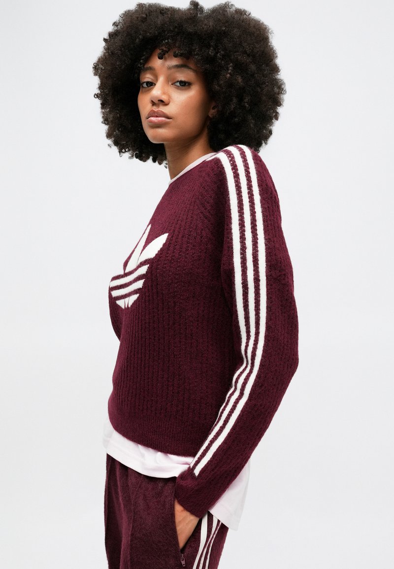 Maglione oversize in bordeaux realizzato a maglia, con strisce bianche adidas lungo le maniche e logo sul petto. Ha una vestibilità comoda e una texture a costine.