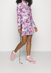Lättviktig golfdräkt med en långärmad topp i rosa och blå blommönster och en matchande kjol, kompletterad med rosa sportskor.
