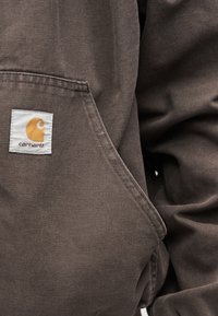 Καφέ ύφασμα Carhartt με ραμμένη μπαλώμα λογότυπου κοντά στην μπροστινή τσέπη ενός ανθεκτικού, υφασμάτινου μπουφάν ή φούτερ με κουκούλα.