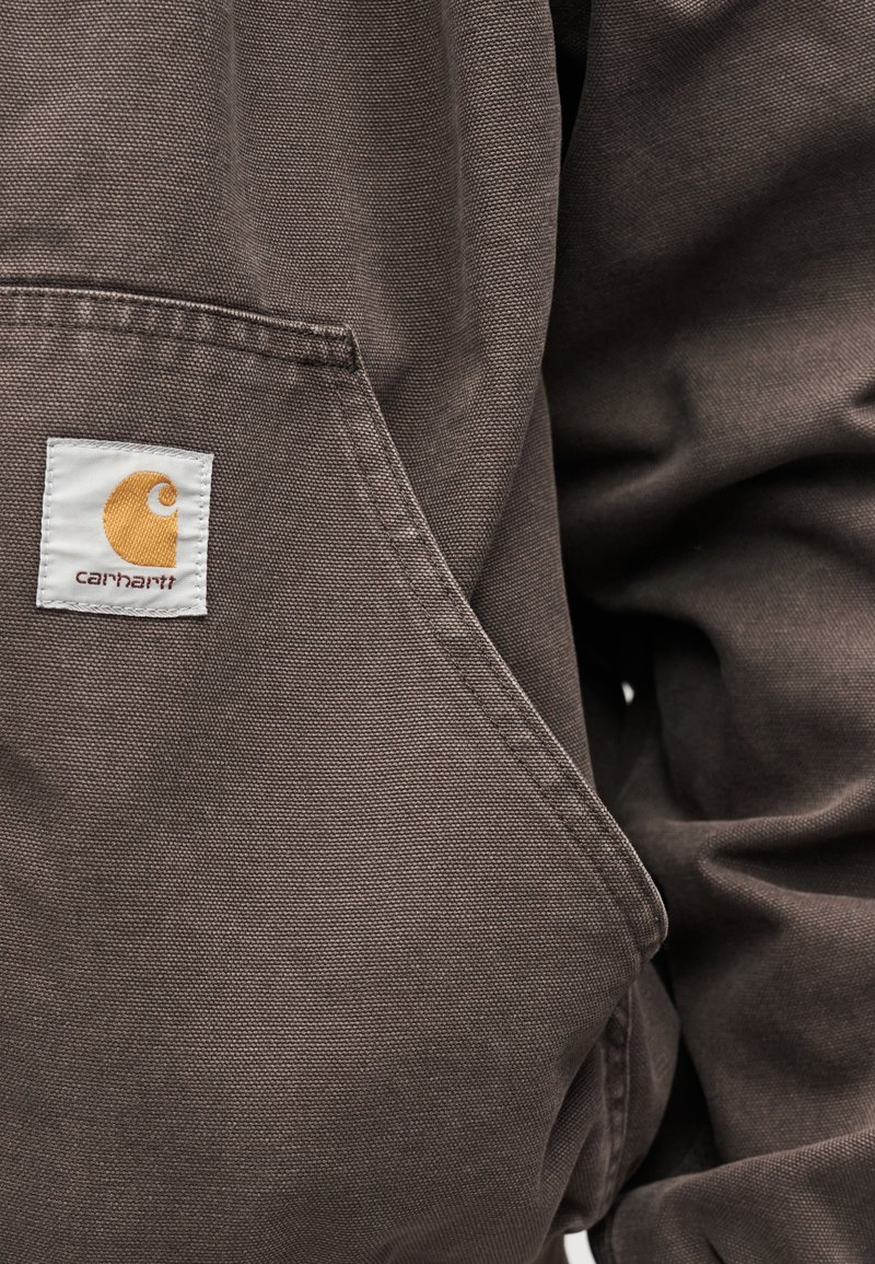 Tessuto Carhartt marrone con logo a patch cucito vicino alla tasca frontale di una giacca o felpa resistente e strutturata.