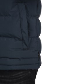 Tamno plavi puffer prsluk s patentnim zatvaračem, teksturiranom površinom i šivanjem, s crnom etiketom na rubu.