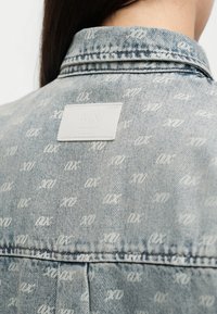Πλάτη ατόμου που φοράει ανοιχτό μπλε τζιν μπουφάν με λευκή εκτύπωση μονόγραμμα και λευκή ορθογώνια ετικέτα στο πάνω μέρος της πλάτης.
