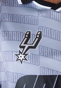 Grijs sportshirt met een groot zwart logo met een ster en de tekst "San Antonio" in een herhalend patroon. Kraag met zwart-witte strepen.