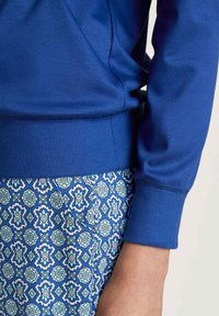Blauwe sweatshirt met geribde manchetten, gecombineerd met gestreepte blauwe en lichtblauwe broek met geometrische ontwerpen. Gladde stoftextuur zichtbaar.