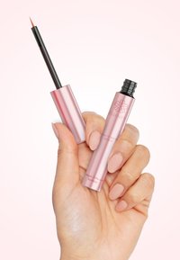 Pink metallic eyelash and brow serum tube held by a hand, featuring a pointed applicator and brand logo on the body. Smooth finish.
