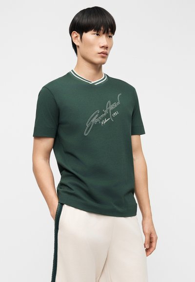 Πράσινο βαμβακερό T-shirt με στρογγυλή λαιμόκοψη, διακοσμημένο με αντίθετες λευκές ρίγες και κεντημένο κείμενο στην μπροστινή πλευρά. Κοντά μανίκια.