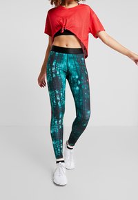 Czerwony, perforowany top i czarny biustonosz sportowy, zestawione z legginsami w tealistym i czarnym wzorze, przedstawiającymi leśne motywy. Całość dopełniają białe buty sportowe.