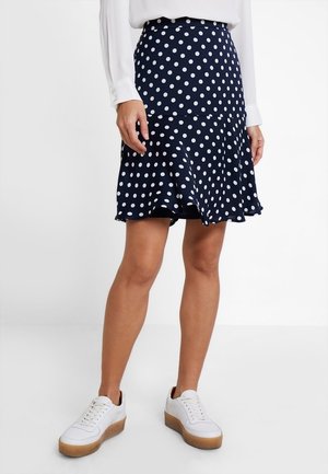 Vrouw met een marineblauwe polkadot rok en witte sneakers, staand met ontspannen handen langs het lijf tegen een effen achtergrond.
