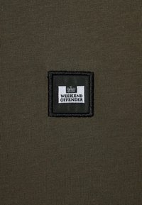 Material de culoare verde măsliniu cu textură canelată, având un patch dreptunghiular negru cu textul "WEEKEND OFFENDER" în alb și un logo cu o coroană.