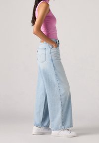 Kobieta w jasnoniebieskich szerokich spodniach, różowej, bezrękawnikowej bluzce i białych sneakersach stoi z rękami w kieszeniach na jednolitym tle.