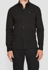 Chemise noire à manches longues avec boutons, fabriquée en tissu lisse, comportant un petit logo blanc sur la poitrine gauche et un col classique.