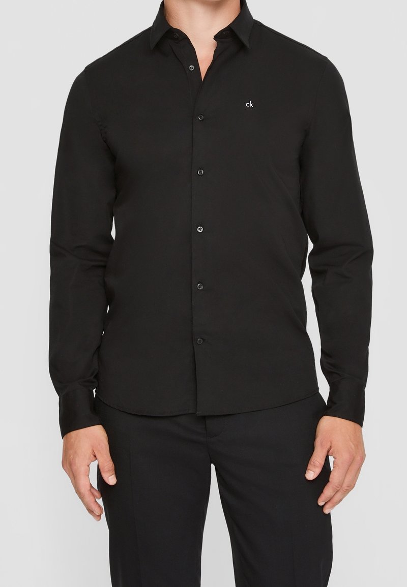 Chemise noire à manches longues avec boutons, fabriquée en tissu lisse, comportant un petit logo blanc sur la poitrine gauche et un col classique.
